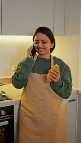 Ein-Glückliches-Brünettes-Mädchen-Mit-Brauner-Haut-In-Einer-Grünen-Jacke-Und-Einer-Beigen-Schürze-Telefoniert,-Während-Es-In-Einer-Modernen-Wohnung-In-Der-Küche-Einen-Snack-Mit-Einem-Croissant-In-Der-Hand-Isst